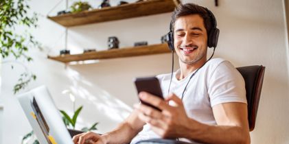 headphone wearing guy sitting in chair looking at phone whle tapping on laptop